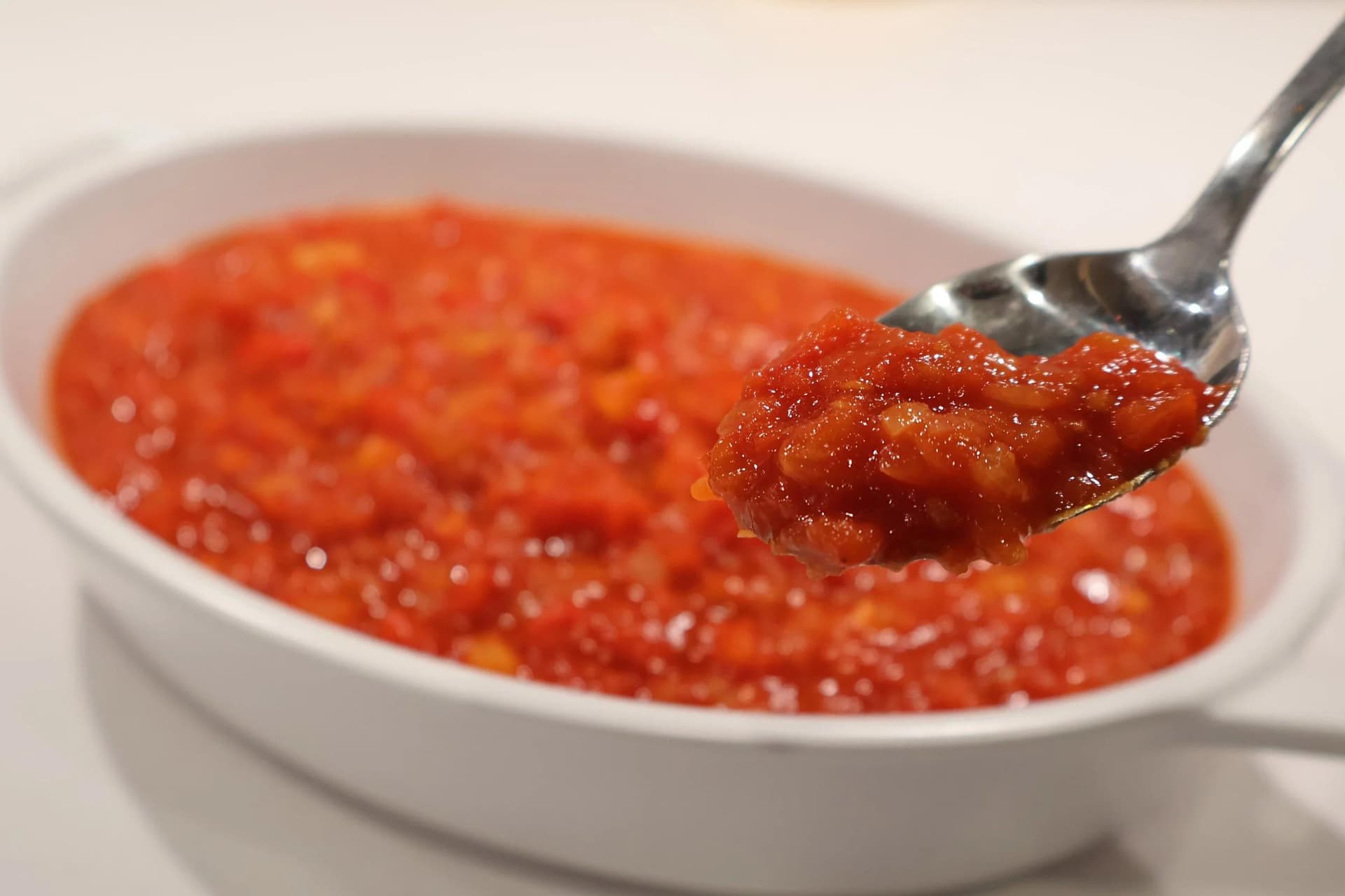 Chunky piperade sauce with visible tomato and pepper pieces in a white oval dish, spooned up to show texture