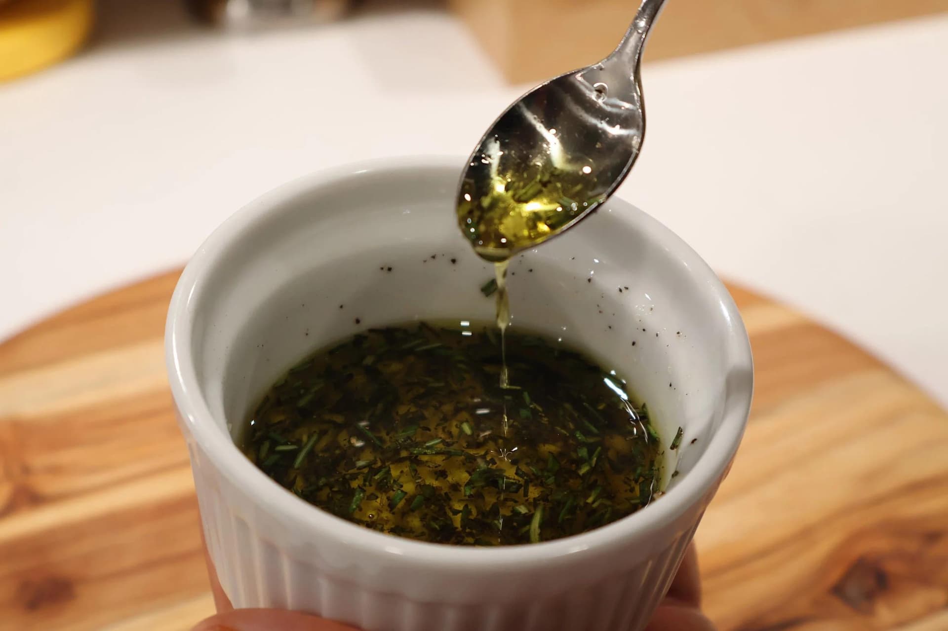 Herb-infused olive oil dripping from a spoon into a white bowl containing green herbs and oil.